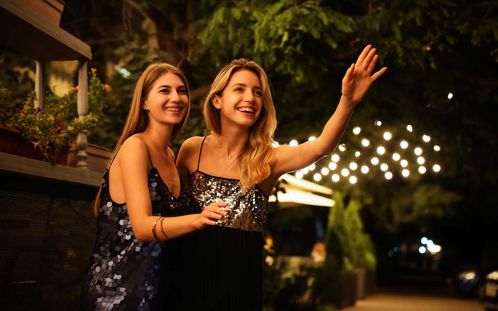 Zwei junge, lächelnde Mädchen stehen umarmt vor einem Lokal am Abend und rufen ein Taxi