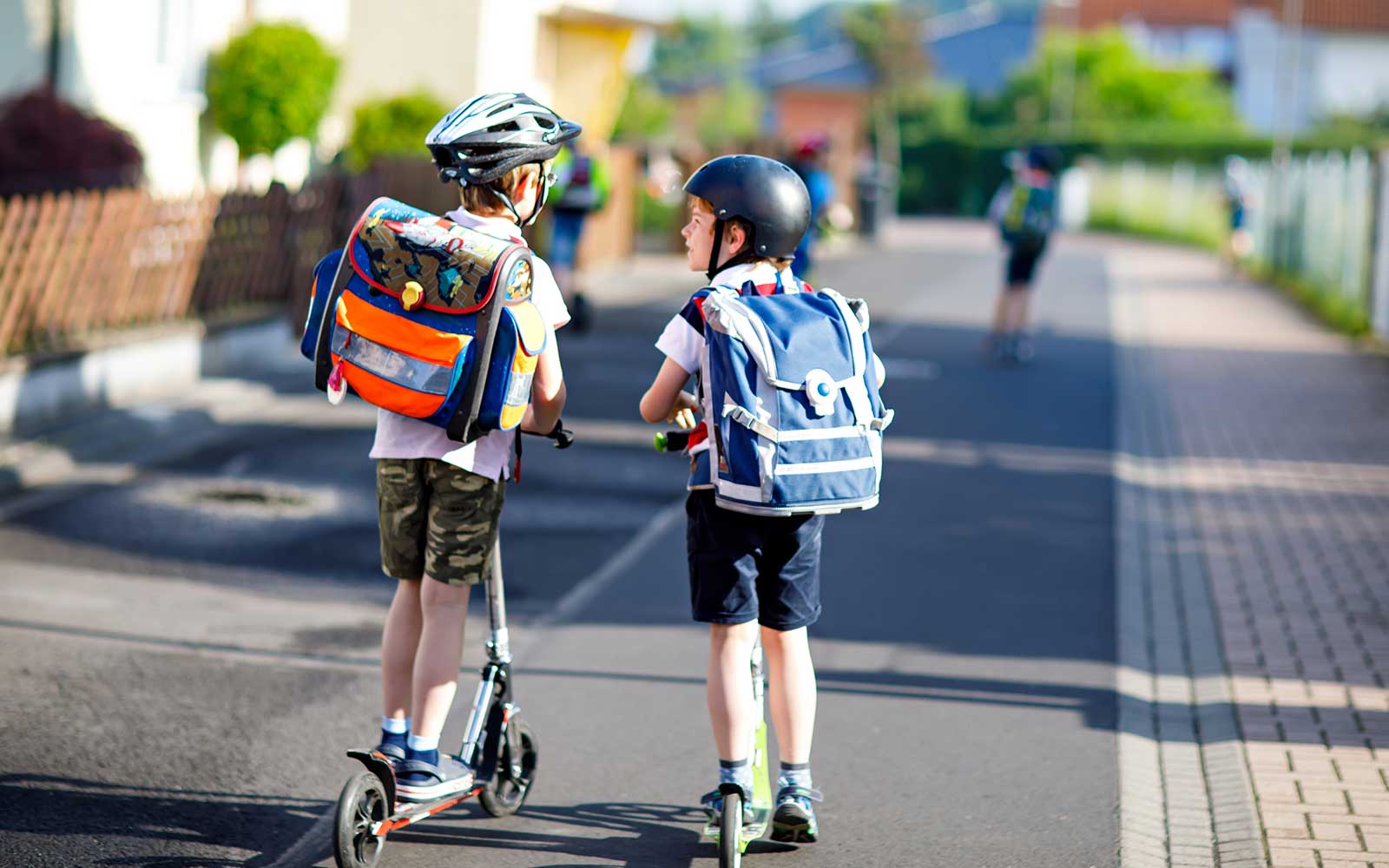 Zwei Schulkinder mit Helmen und Schultasche fahren auf ihren Scooters eine Straße entlang